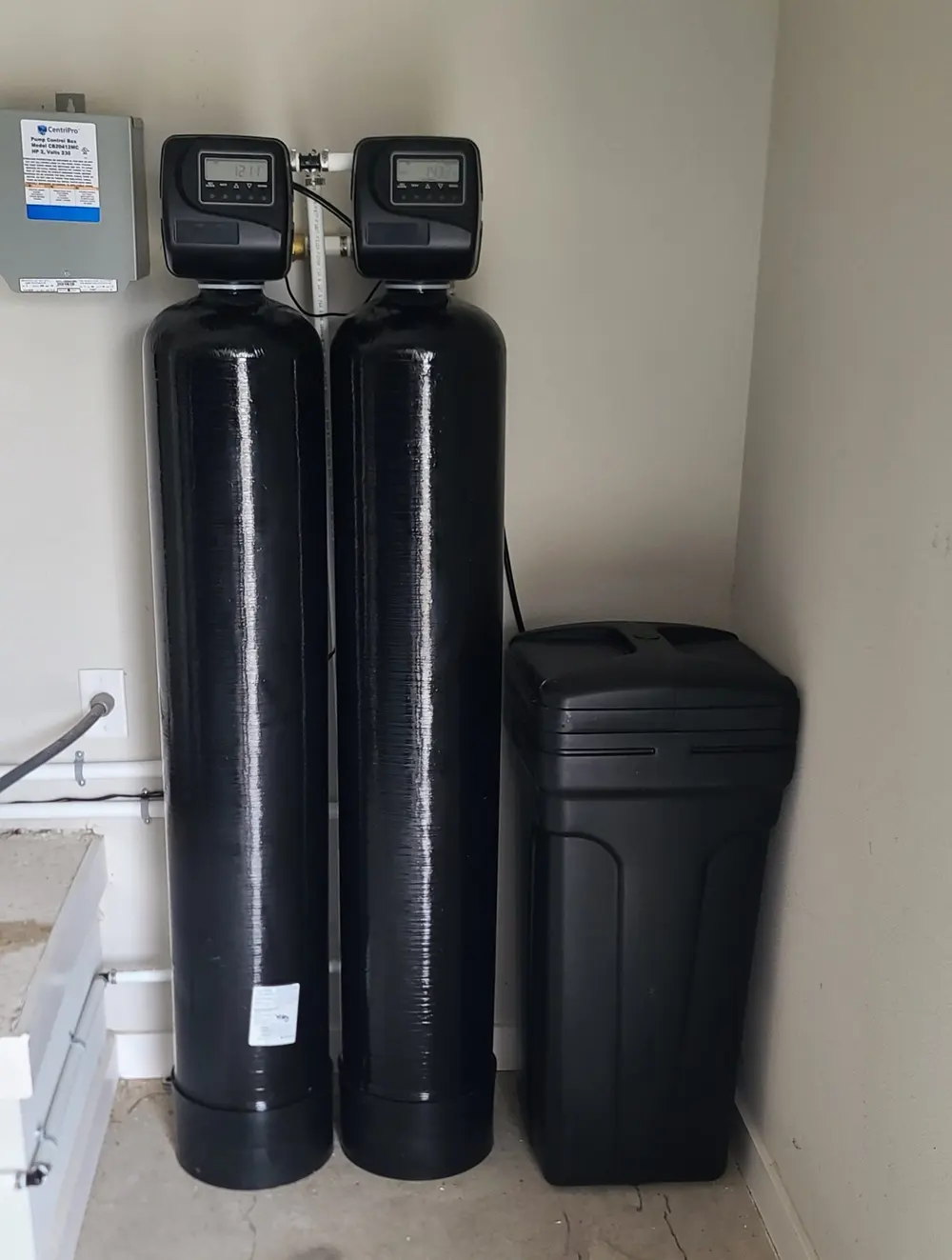 Twin metered tanks and brine tank in a utility corner alongside a well pump control box